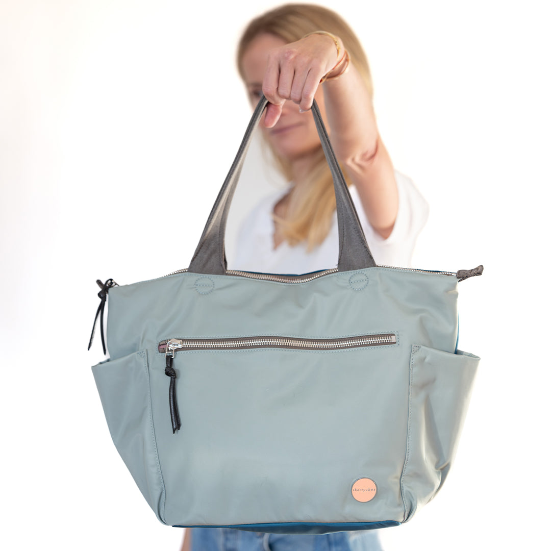 Person holding a lslate/teal tillie tote bag with a visible brand logo on a white background