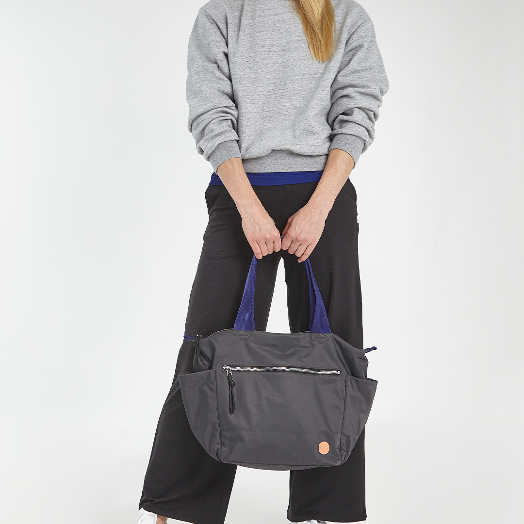 Person holding a charcoal gray tillie tote bag with a visible brand logo on a white background