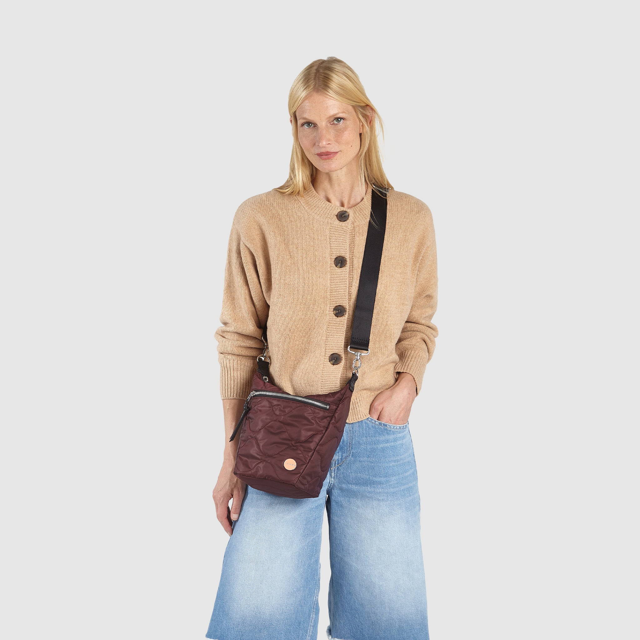 Woman wearing a beige cardigan and blue jeans, holding a bordeaux marcel quilted crossbody  bag against a white background